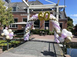 Ballonboog foliecijfers 60 met gouden, witte en lila ballonnen en Ballontrossen bij brug